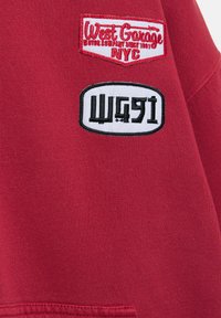 Rødt stof med to påsyede patches: øverst i rød og hvid står der "West Garage NYC," nederst i sort og hvid vises det stiliserede "WG91".