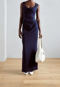 Marineblauw lang jurkje met kantaccenten, gedrapeerd ontwerp en mouwloze uitsnijding. Gecombineerd met een witte handtas en wijde witte schoenen op een houten vloer.