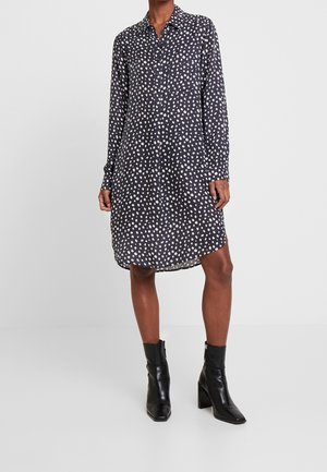 Robe chemise noire à manches longues avec des points blancs irréguliers, longueur genou, associée à des bottines à talons noires sur un modèle à la peau de teinte moyenne foncée.