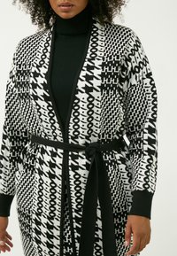Cardigan a righe bianche e nere con design geometrici, cintura da annodare e polsini a costine, abbinato a un dolcevita nero sotto.