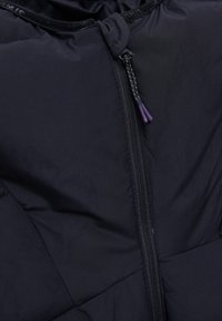 Gros plan sur la fermeture éclair noire d'une veste avec deux cordons de tirage à pointes violettes et des panneaux de tissu cousus.