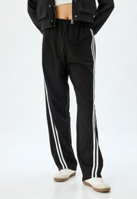 Schwarze Sporthosen mit zwei weißen Seitenstreifen, aus glattem Stoff, mit einem elastischen Bund und lockerer Passform.