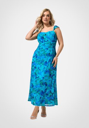 Blondynka ubrana w niebieską, kwiecistą, bezrękawnikową sukienkę maxi i srebrne szpilki, stojąca z jedną ręką przy włosach, drugą wzdłuż boku.