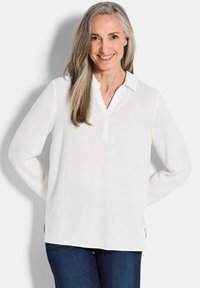 Blusa bianca a maniche lunghe con colletto e vestibilità leggermente rilassata, completata da fessure laterali per facilità di movimento.