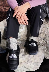 Mustat pehmeät slip-on-kengät, joissa on valkoinen logo, käytetty harmaiden neulos sukkien päällä. Asetettu kivipinnalle, yhdistetty mustiin housuihin ja vaaleanpunaiseen toppiin.