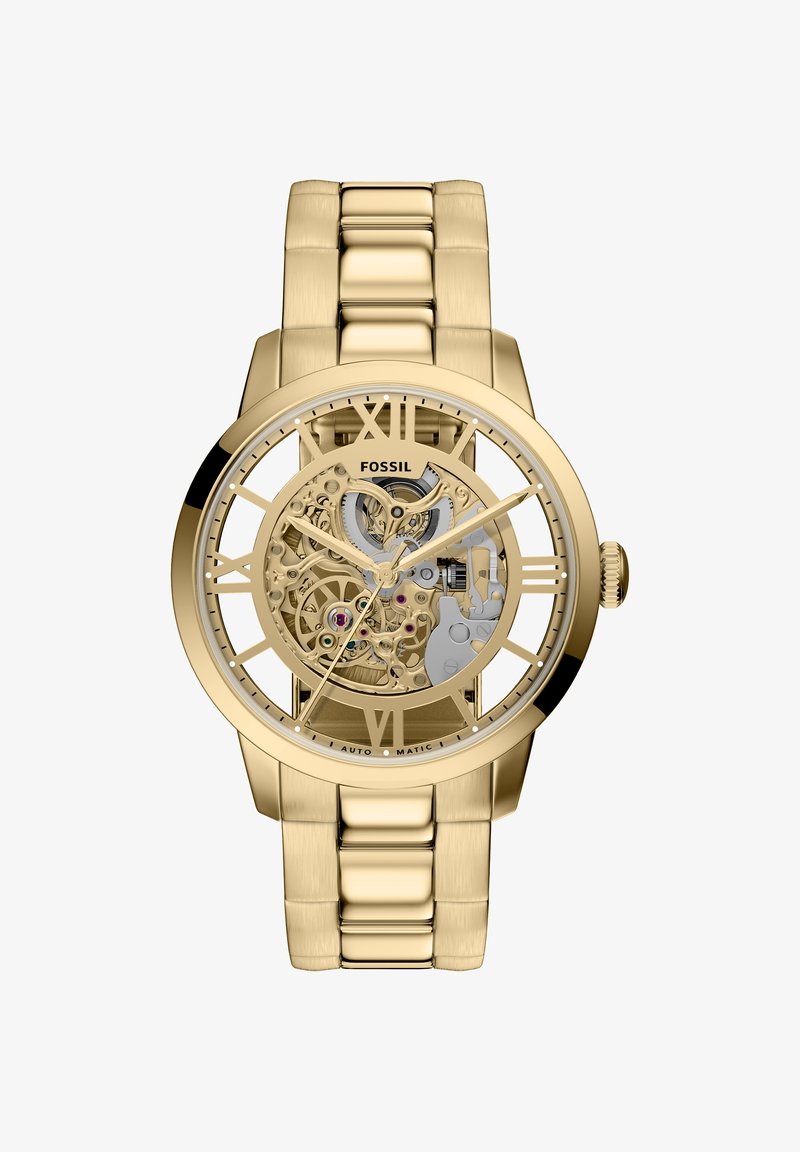 Montre squelette ton or avec chiffres romains, bracelet à maillons en acier inoxydable, détails du mouvement visibles et finition polie.