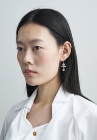 Sudraba auskari kā piliens ar sarežģītiem ģeometriskiem rakstiem, kuros ir zvaigžņu formas. Modelis valkā baltu kreklu ar nelielu apkakli.