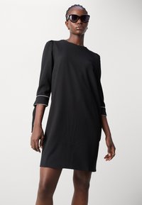 Black dress with three-quarter sleeves and white accent at cuffs, featuring a straight cut and smooth texture. Worn with oversized black sunglasses.