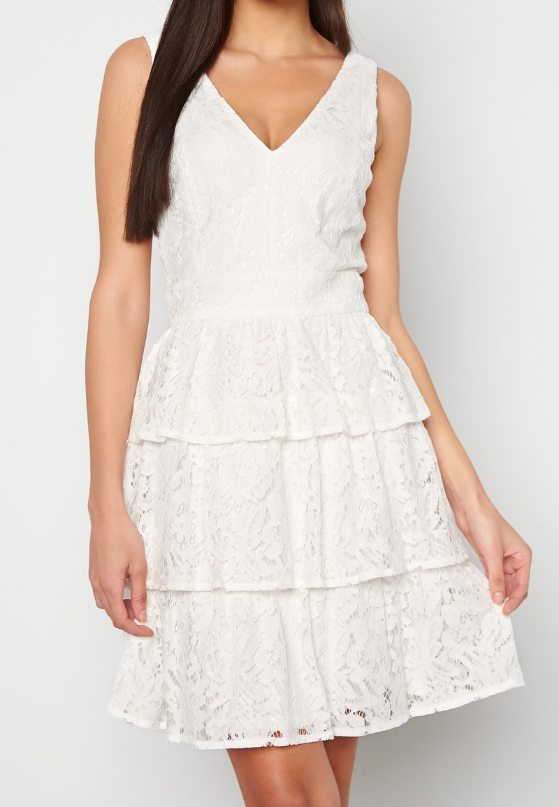 Abito bianco in pizzo con scollatura a V, volant a strati e corpetto aderente. Il tessuto presenta motivi floreali e un dettaglio testurizzato.