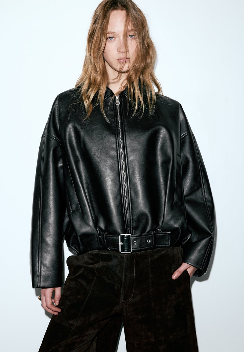 Fekete bőr túlméretezett cipzáras dzseki öves derékrésszel, sima textúrával és minimalista dizájnnal, sötétbarna velúr nadrággal párosítva.