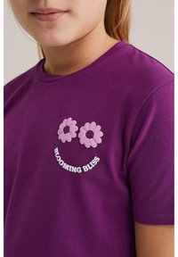 Camiseta de algodón morada con mangas cortas, decorada con apliques de flores rosas y el texto "BLOOMING BLISS" en blanco, ubicado cerca del escote.