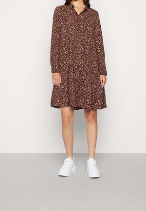 Vrouw draagt een bruine jurk met kraag, roze stippen en lange mouwen, gecombineerd met witte sneakers, staand tegen een effen achtergrond.