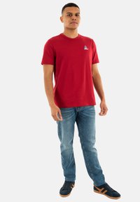 Camiseta de algodón roja con cuello redondo, que presenta un pequeño gráfico en el pecho. Combinada con jeans de color azul claro y zapatillas negras con detalles marrones.