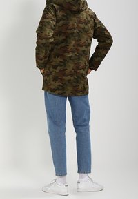 Kamo parka s kapucí, zdobená kombinací zelených a hnědých vzorů. Nosená s světle modrými tapered džínami a bílými teniskami.