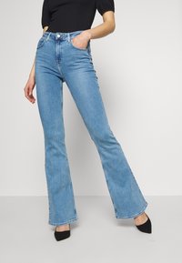 Frau in hellblauen, hochgeschnittenen Schlagjeans, schwarzem Top und schwarzen, spitzen High Heels, mit einer Hand in der Tasche, vor einem schlichten weißen Hintergrund stehend.