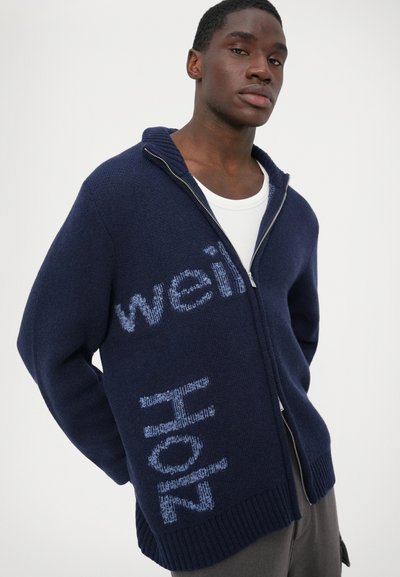 Marinblå stickad zip-tröja med ribbad nederkant. Har ett ljusblått textmönster "vi" och "heta" på framsidan. Mjuk textur, avslappnad passform.
