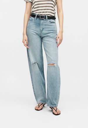 Γυναίκα που φοράει ανοιχτό μπλε τζιν παντελόνι με σκισίματα και φαρδιά μπατζάκια, μαύρη ζώνη, ριγέ κοντομάνικη μπλούζα και μαύρα σανδάλια με τακούνι σε λευκό φόντο.