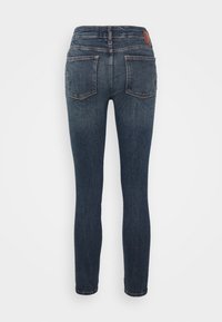 Jeans ajustados de mezclilla azul oscuro con detalles desgastados, cinco bolsillos y una parche de cuero marrón en la cinturilla trasera.