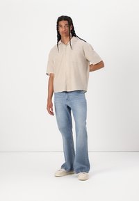 Chemise beige à manches courtes avec une poche poitrine, associée à un jean évasé bleu clair et des baskets de couleur crème sur un fond blanc.