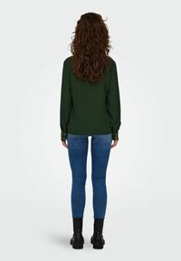Grøn langærmet bluse med knappede manchetter, parret med tætsiddende blå jeans og sorte ankelstøvler. Bagudblik, der viser tekstureret hår.