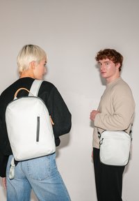 Zwei Personen zeigen Taschen: einen hellgrauen Rucksack mit einem Reißverschlussfach und braunen Akzenten sowie eine kleine graue Umhängetasche mit einem schwarzen Rand.