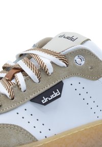 White leather sneakers with beige suede accents, brown and white striped laces, perforated sides, and a black logo on the side.