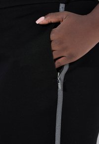 Ciemnoskóra dłoń z jasnoróżowym lakierem na paznokciach spoczywa w kieszeni czarnego ubrania z srebrnym suwakiem.