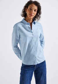 Vrouw met krullend donker haar, draag een paar zilveren creolen, een lichtblauwe gestreepte button-up shirt en een blauwe spijkerbroek, staand met haar handen achter haar rug.