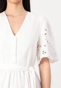 Vit klänning med v-ringning och knappdetaljer. Korta spetsärmar med broderade utskärningar. Knytlinning för ökad definition. Mjuk tygtextur.