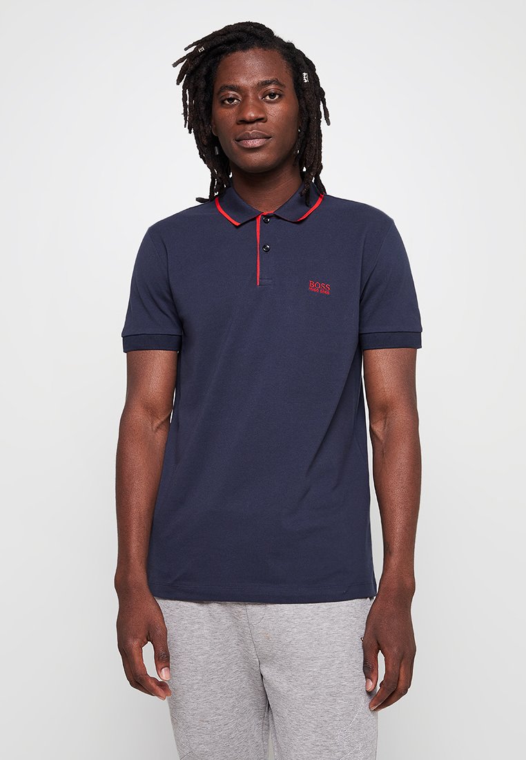 Polo shirt bleu marine avec col et finitions rouges, à manches courtes, et logo brodé sur la poitrine. Matière en coton lisse, coupe classique.