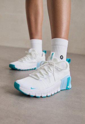Jambes portant des chaussettes blanches et des chaussures de sport blanches avec des accents turquoise, debout sur un sol gris lisse.