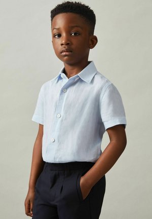Camicia azzurro chiaro a maniche corte con bottoni, realizzata in lino, abbinata a pantaloni neri scuri; la camicia presenta un colletto a punta e una patta frontale.
