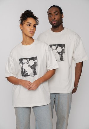 Zwei Erwachsene tragen weiße, übergroße T-Shirts mit schwarz-weißen Blumenmustern, beide stehen vor einem schlichten grauen Hintergrund.