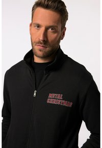 Chaqueta negra con cremallera y cuello alto, que presenta el texto bordado en rojo "METAL CHRISTMAS" en el lado izquierdo del pecho. Textura de tela suave.
