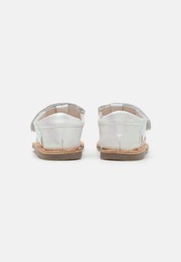 Sandalias de cuero metálico plateado con suela plana, diseño abierto en la parte trasera y correa ajustable. La plantilla es de color marrón claro y la suela exterior es marrón.