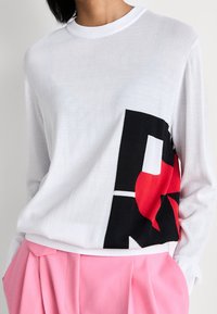 Pull blanc à manches longues avec un motif géométrique noir et rouge sur le côté, associé à un pantalon plissé rose taille haute, mains dans les poches.
