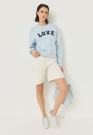 Frau trägt hellblaues Sweatshirt mit dem Schriftzug "LOVE", beige Shorts, weiße Sneakers und hält eine hellblaue Handtasche, steht vor einer weißen Wand.