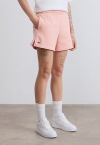 Shorts en coton rose clair avec une taille élastique et des poches latérales. Présentent un petit logo vert sur le côté et une coupe décontractée.