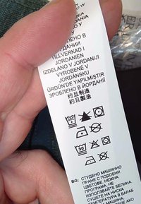 Primer plano de una mano sosteniendo una etiqueta de cuidado de la ropa con símbolos de lavado y texto multilingüe indicando que el artículo está hecho en Jordania.