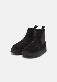 Botas negras hasta el tobillo hechas de suave gamuza con paneles elásticos, una suela de plataforma gruesa y tiradores traseros para ponérselas fácilmente. Textura lisa sin patrones.