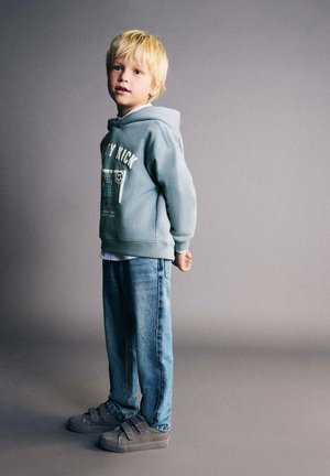 Hanorac albastru deschis cu imprimeu grafic, blugi din denim albastru și adidași gri cu velcro. Păr blond și fundal neutru. Poză în picioare.