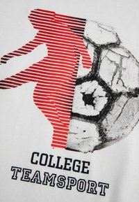 Biała koszulka z czerwoną, prążkowaną sylwetką zawodnika oraz czarno-białą, teksturowaną piłką nożną, z napisem "COLLEGE TEAMSPORT" poniżej.