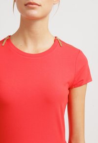 Ljus korall kortärmad t-shirt med rund halsringning och gulddragkedjestängningar på axlarna. Slät bomullsaterial, figurnära design.