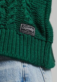 Zielony sweter z dzianiny z prążkowanym hemem; posiada czarną metkę z napisem "Superdry" oraz "Outdoor Outfitters". Tekstura wygląda na grubą.