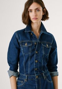 Jeansfarget jumpsuit med mørkeblå vask, med krage, knappelukking foran, to brystlommer, og oppbrettede ermer med kontrastfargede mansjetter.