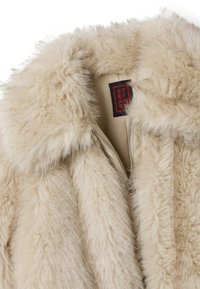 Cappotto in pelliccia sintetica di un beige chiaro con una texture morbida e soffice. Presenta una chiusura con zip e un colletto, con un'etichetta di marca ben visibile.