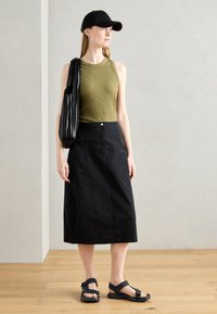Oliven-farget ribbet tank topp, svart kalv-langt skjørt, svart caps, svart bøtteveske og svarte sandaler med justerbare stropper. Bakgrunn med tregulv.