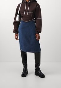 Brun kort hoodie med vit sömnad, kombinerad med en knappad blå denimkjol och knähöga svarta stövlar. Släta texturer och kontrasterande färger.