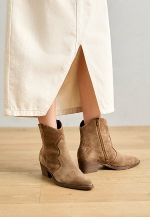 Bottines en suède marron avec fermeture éclair, bout pointu et coutures décoratives en zigzag sur les côtés. Sur un sol en bois clair.
