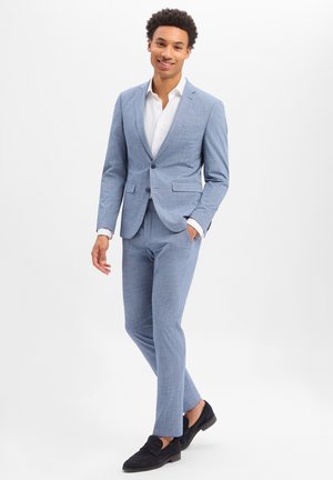 Jeune homme souriant, portant un costume bleu clair avec une chemise blanche et des mocassins noirs, posant devant un fond gris clair.